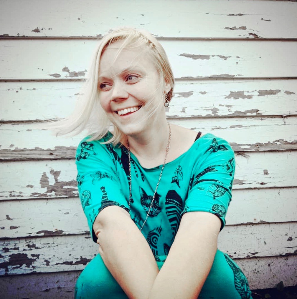 Tiina, a blond woman, pictured against a white wooden wall, looking off-camera and smiling, wearing a turquoise dress.
